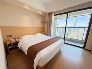Een hotelkamer met een groot bed, een nachtkastje, een lamp en uitzicht op een stadsgezicht door een groot raam.