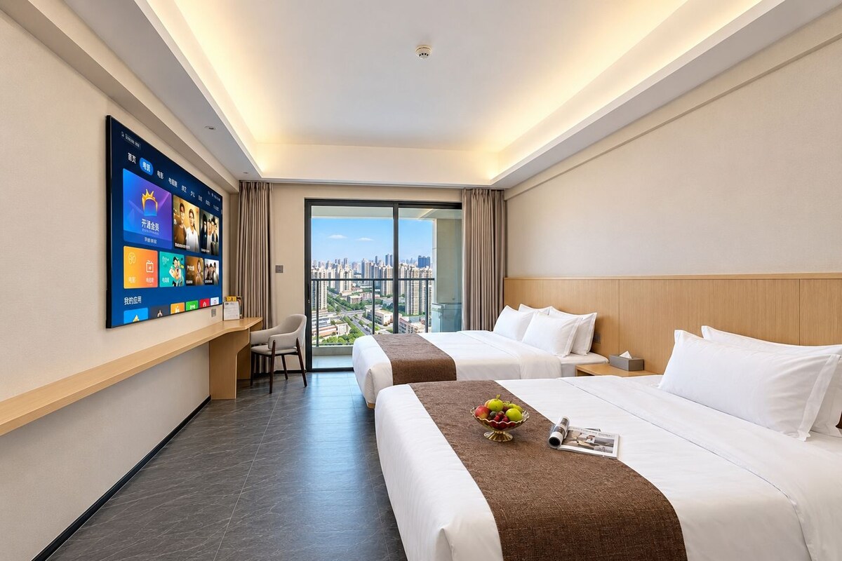 A hotel room with two beds, a desk, a TV mounted on the wall, and a view of the city through the window.