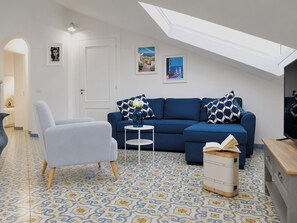A modern living room with a blue sofa, a light grey armchair, a small white side table, and a large window with a skylight. - New Listing (Napoli)
