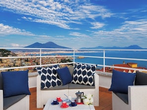 A balcony with a white wicker sofa, two blue cushions, a table with cups and a coffee pot, and a scenic view of a coastal city with mountains in the background. - New Listing (Napoli)