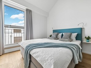 Ein Schlafzimmer mit einem Bett, einem Fenster mit Vorhängen, Blick auf Gebäude und einem türkisblauen Kopfteil.
