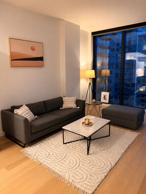 Ein modernes Wohnzimmer mit einer Couch, einem Couchtisch und einer Lampe. Ein großes Fenster bietet Blick auf Gebäude.