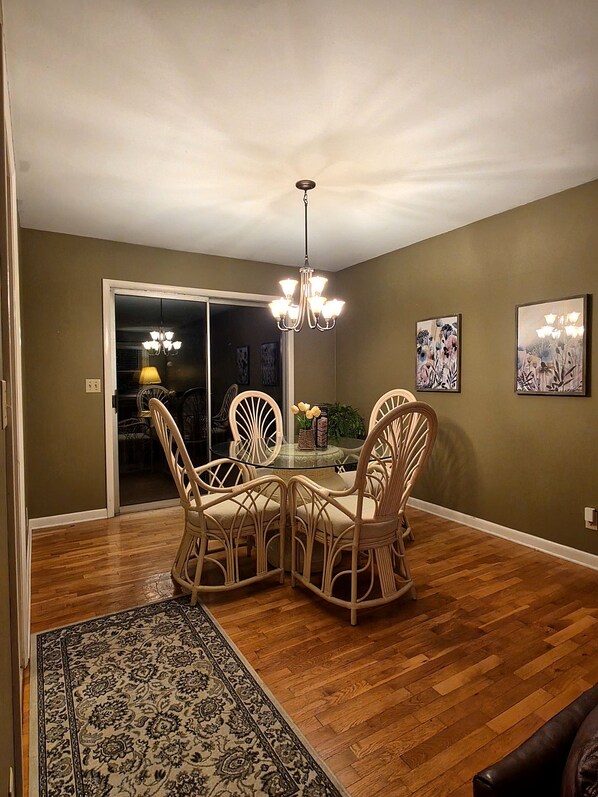 A dining area with a glass-top table surrounded by wicker chairs, a chandelier, and a patterned rug.