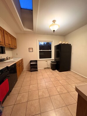 A kitchen with a window, a black refrigerator, wooden cabinets, a dishwasher, and a trash can.