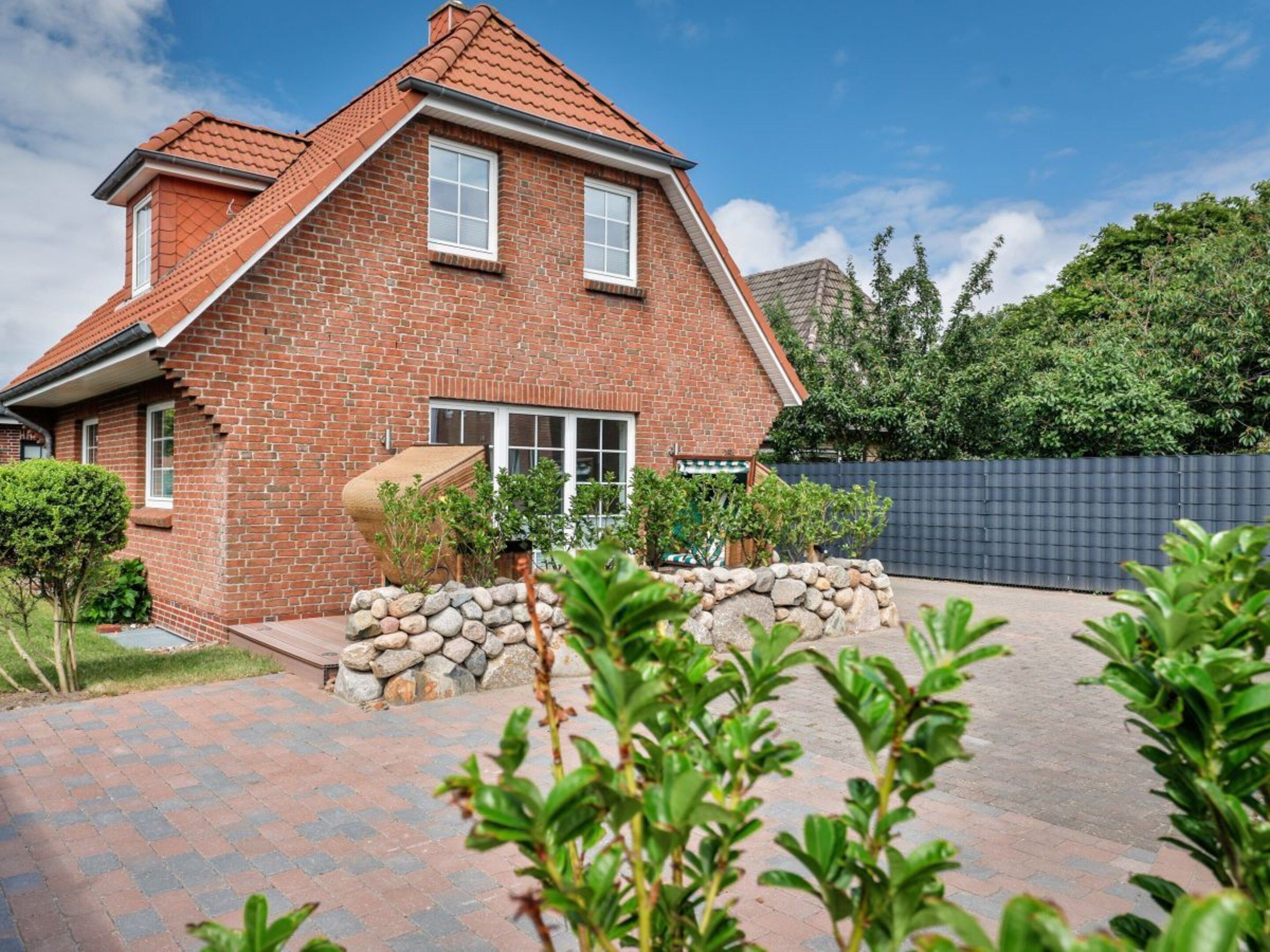 A two-story brick house with a sloped roof, a stone wall in front, and a paved driveway.