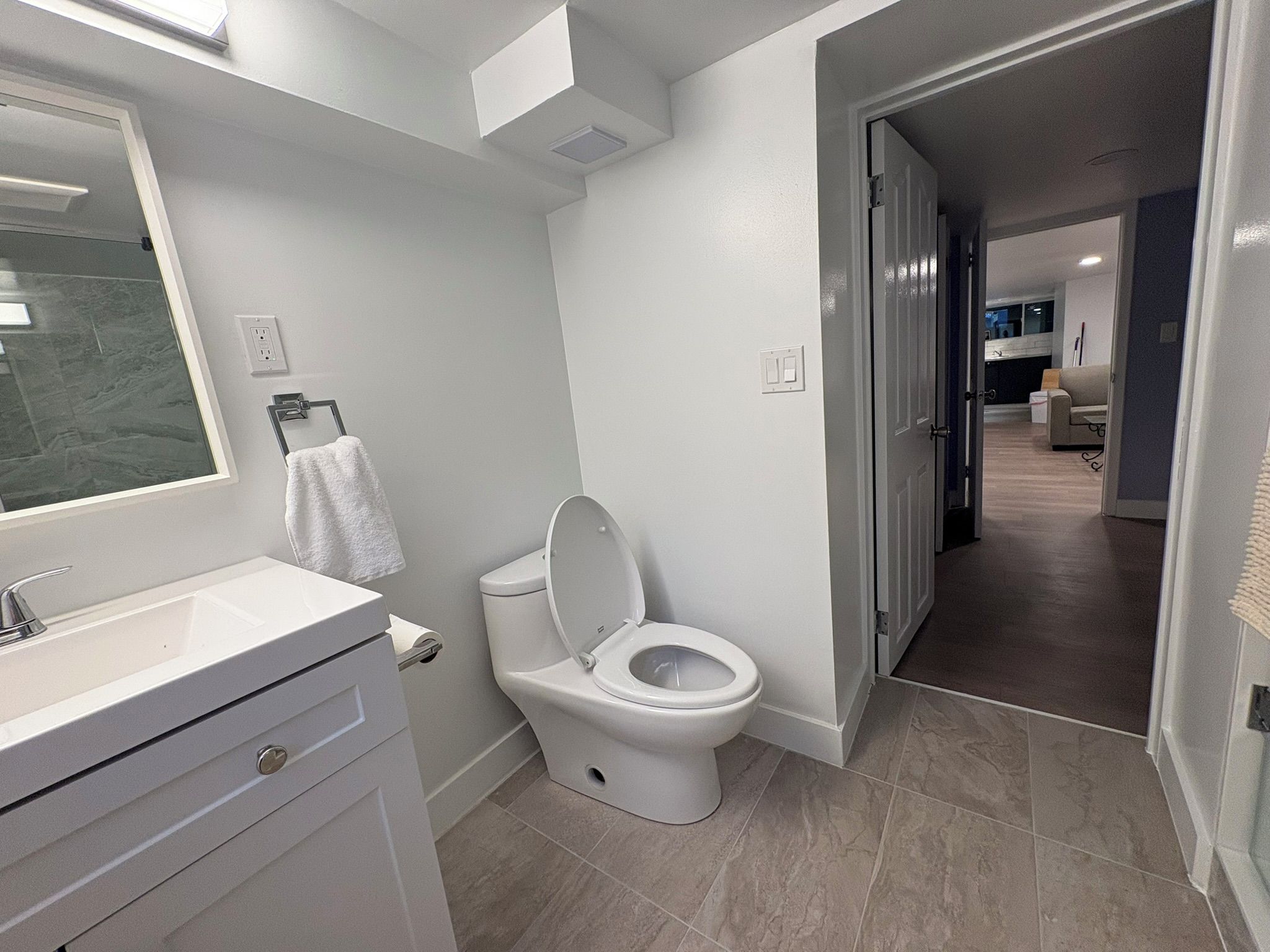 Une salle de bain moderne avec un lavabo, des toilettes et un miroir, tous de couleur blanche. Une serviette est suspendue sur un porte-serviettes, et le sol est carrelé.