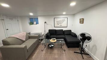 A modern living room with a sofa, a smaller couch, a glass coffee table, a radiator, and a fan.
