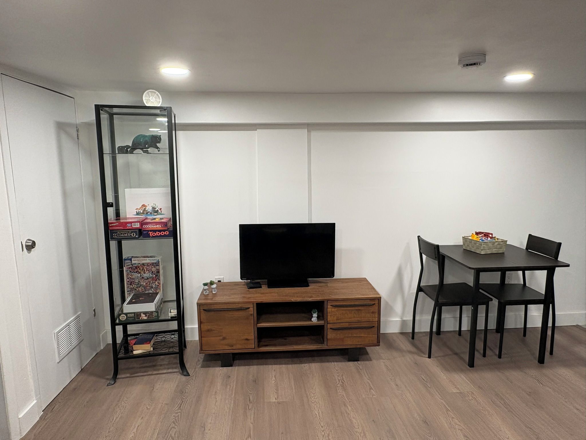 A room with a wooden entertainment unit, a television, a glass display cabinet, and a dining table with chairs.