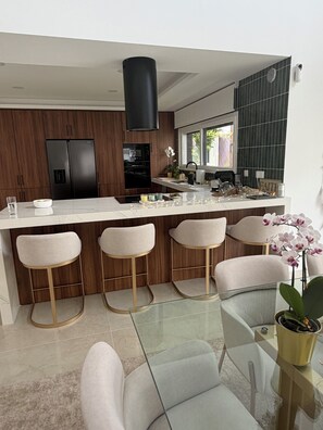 A modern kitchen with a central island, bar stools, and a glass-top dining table. - Luxury New Lisbon/Beach Villa-3 bd, 4 bth, Jacuzzi, Fireplaces, Heated Salt Pool (Setúbal)