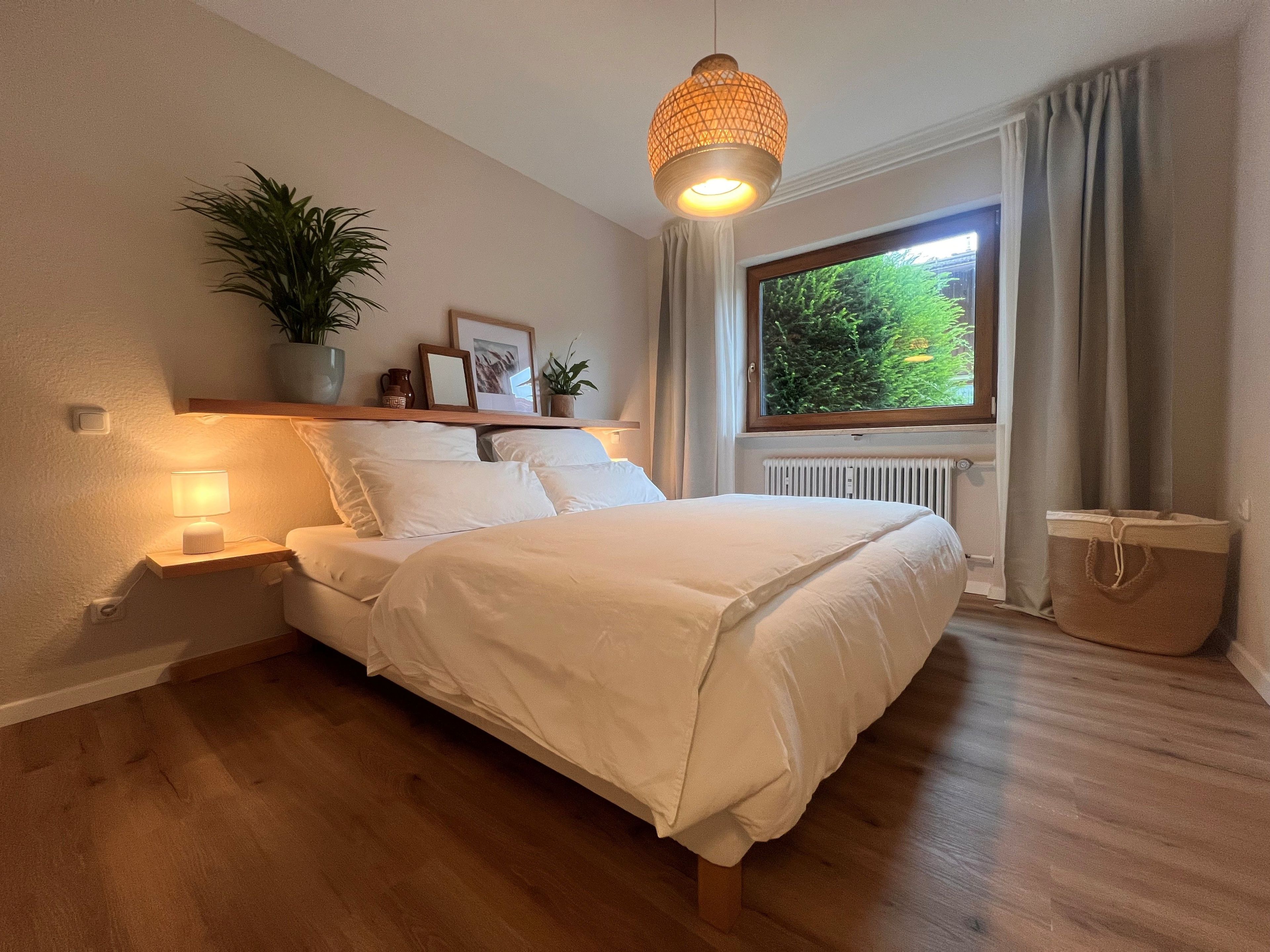 A bedroom with a bed, bedside lamps, a window with curtains, a radiator, and a potted plant.