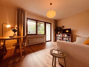 A cozy living room with a sofa, a coffee table, a bookshelf, a window with curtains, and a radiator. - Small, cozy, charming—a bright home in Chiemgau (Ruhpolding)