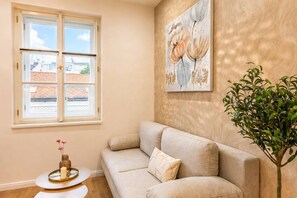 Una sala de estar con un sofá color beige, una mesita redonda con una vela, una planta en maceta y un ventanal con vistas a edificios.
