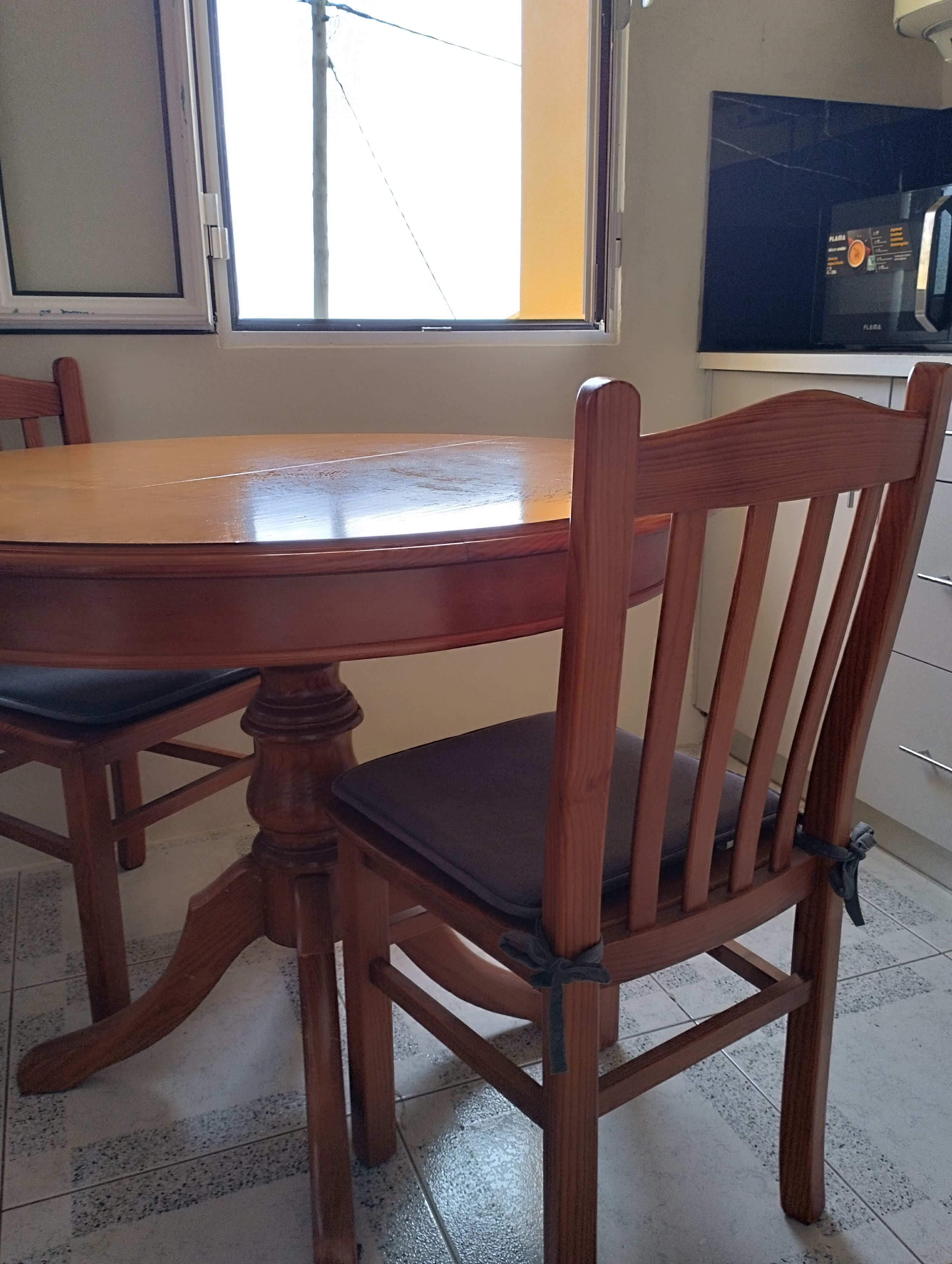 Een houten eettafel met stoelen, een raam met zonwering, en een keuken met kasten.