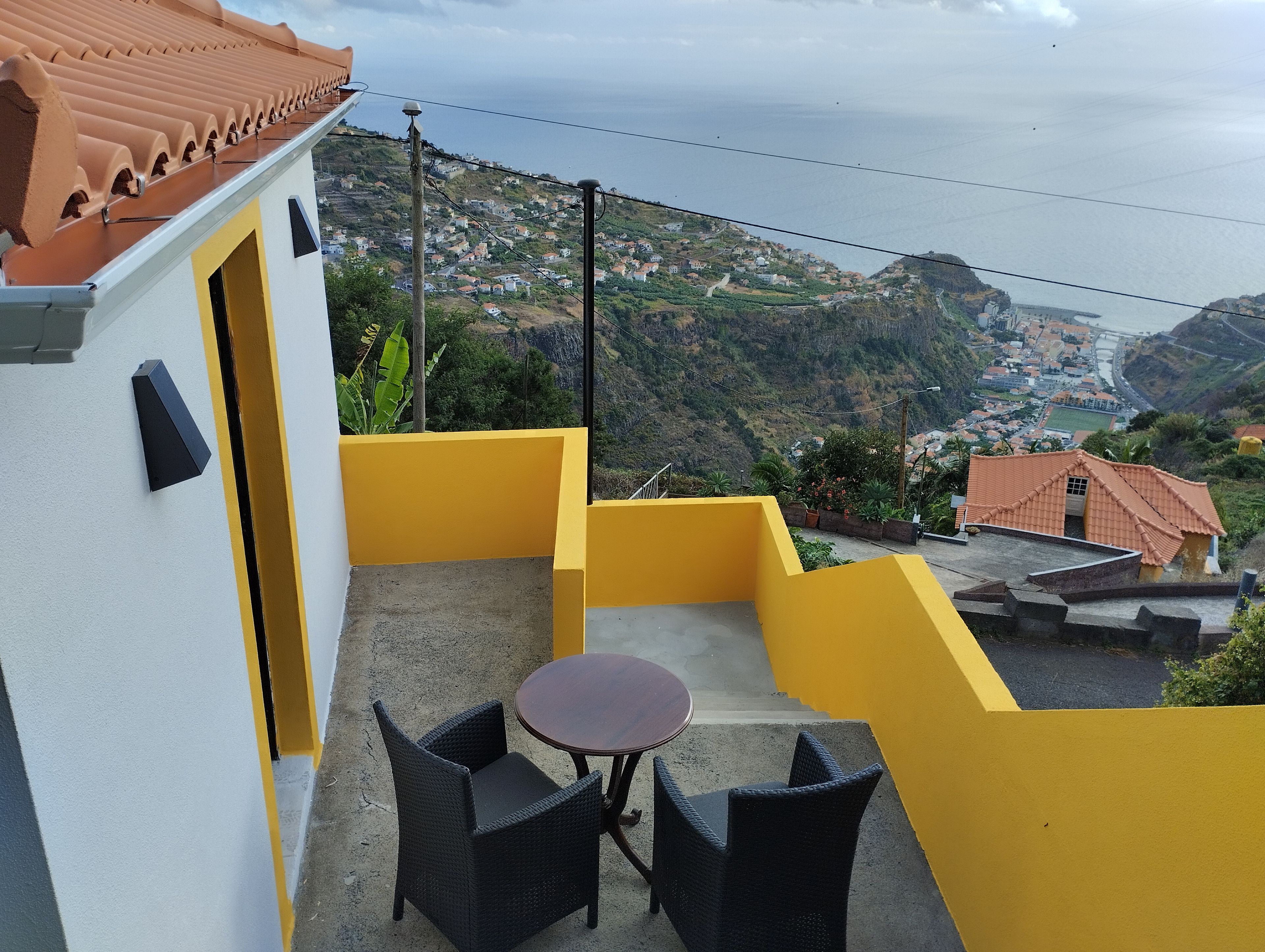 Un balcón con dos sillas de mimbre y una mesita redonda, con vistas a un pueblo costero con casas escalonadas y al mar.