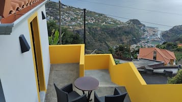 Un balcón con dos sillas de mimbre y una mesita redonda, con vistas a un pueblo costero con casas escalonadas y al mar.