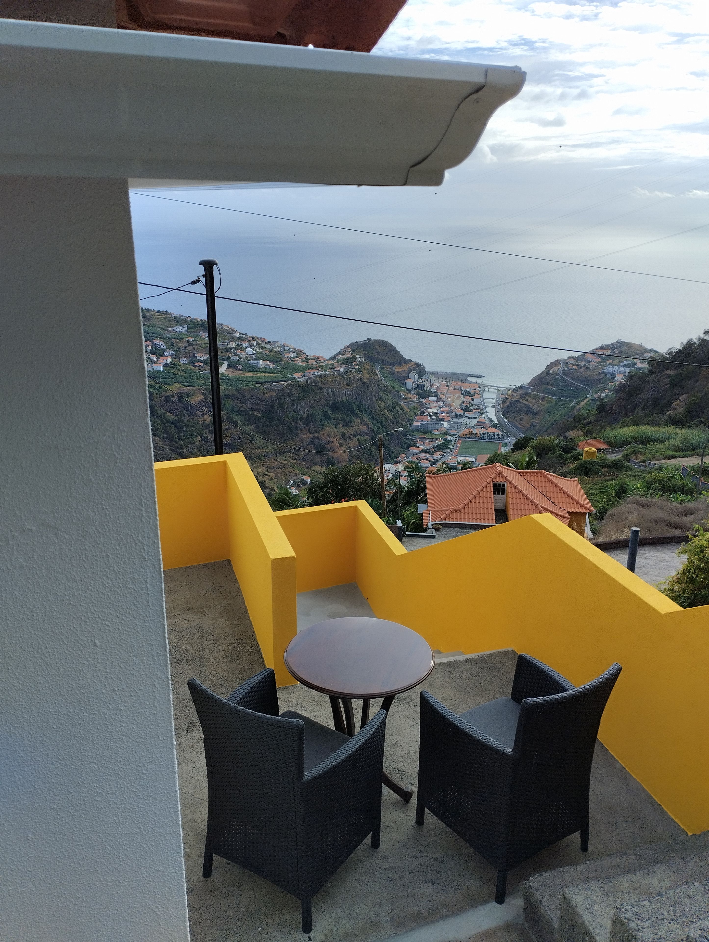 Un balcon avec une table et des chaises, donnant sur une ville côtière avec ses maisons et un paysage vallonné.