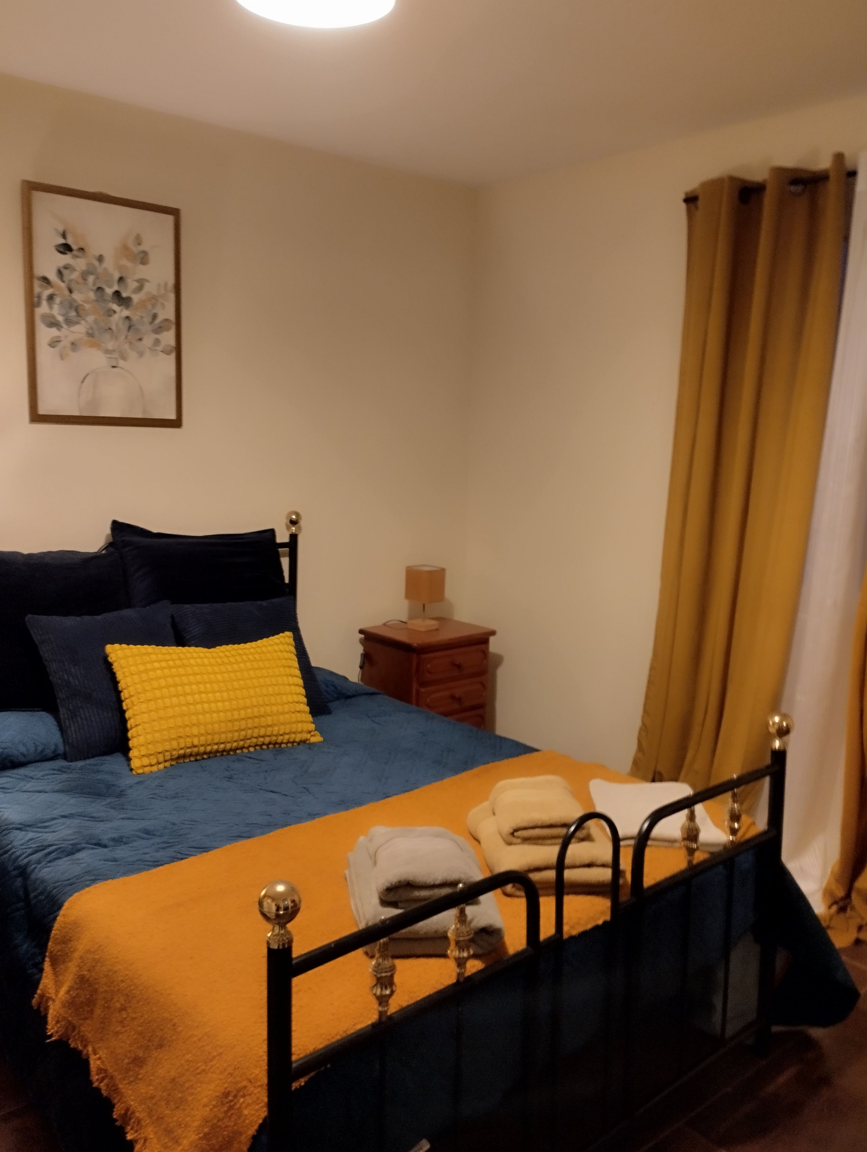 Een slaapkamer met een bed, nachtkastje, lamp en een schilderij aan de muur.