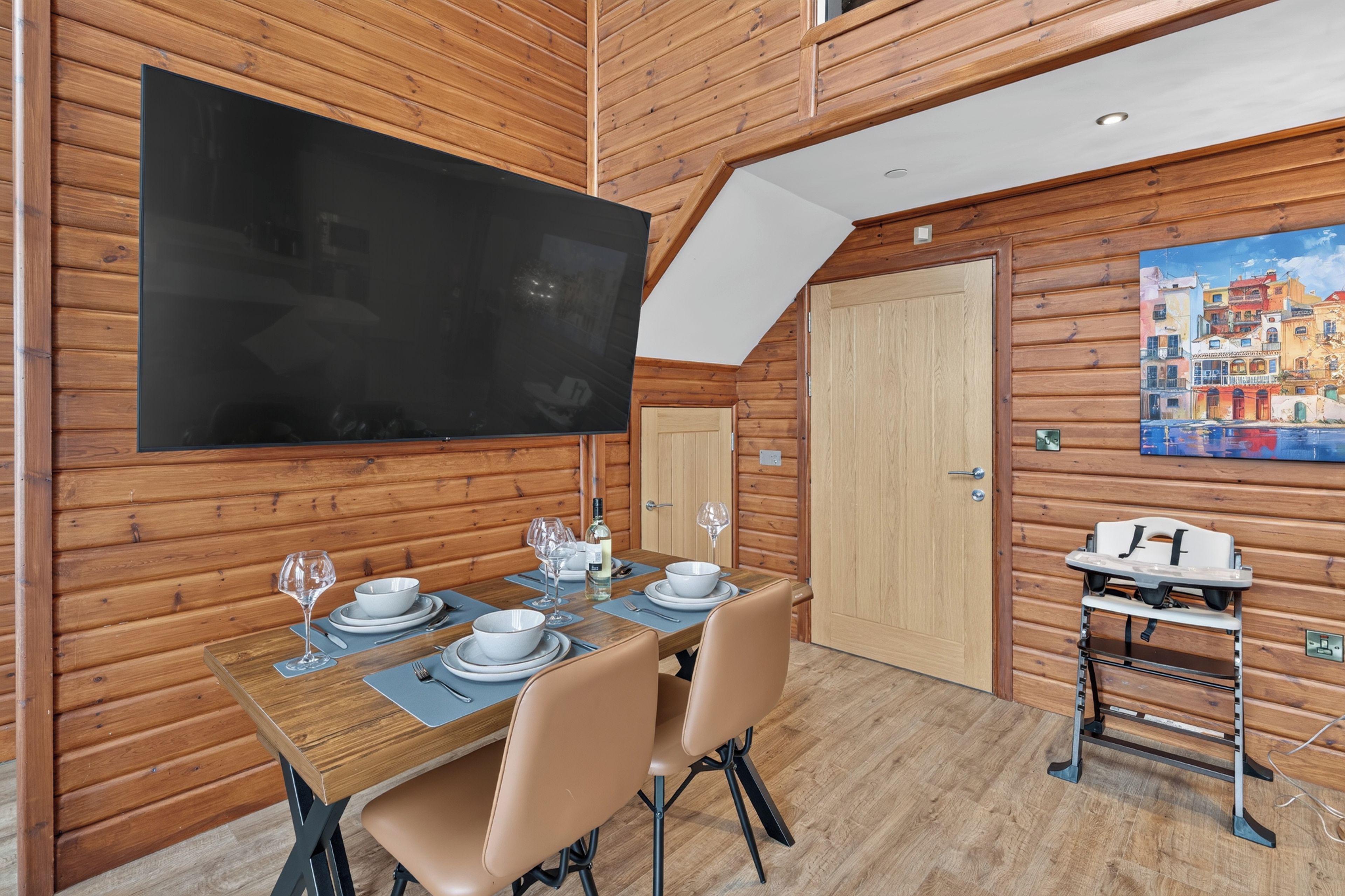 A dining area with a wooden table set for two, a television mounted on the wall, and a painting of a coastal scene.