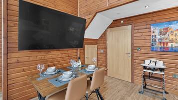 A dining area with a wooden table set for two, a television mounted on the wall, and a painting of a coastal scene.