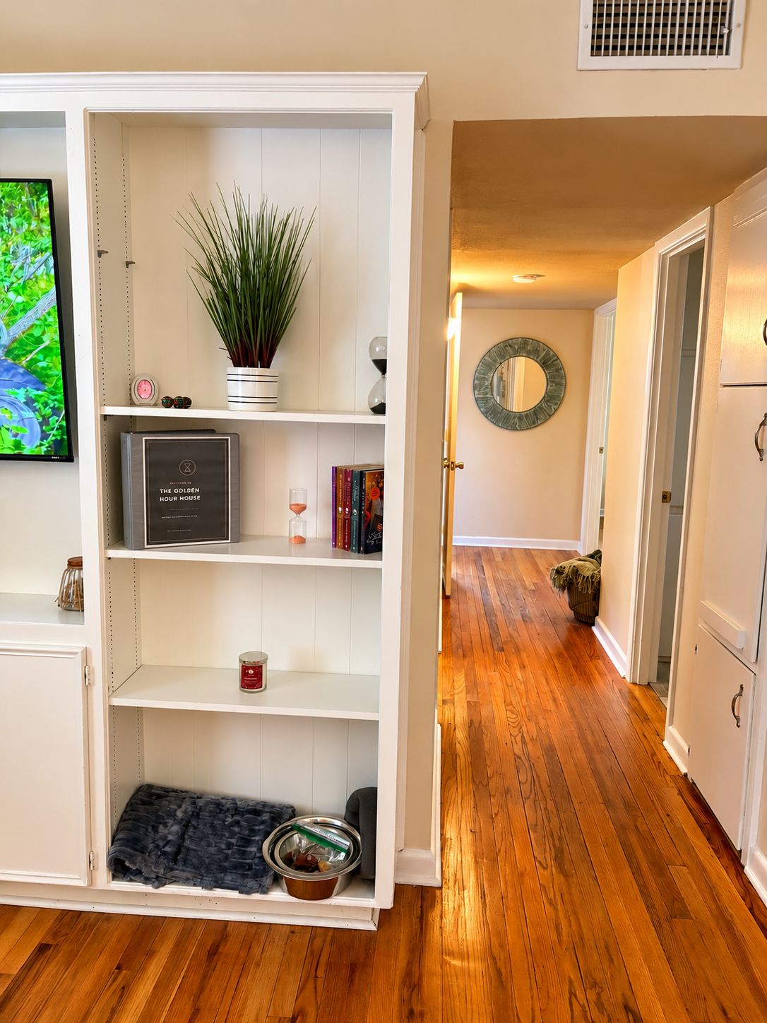A room with a white built-in shelving unit, a potted plant, a clock, a framed mirror, and a wooden floor.