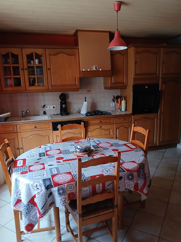 Una cocina con armarios de madera, salpicadero de azulejos, una mesa redonda con mantel acolchado y una lámpara colgante.