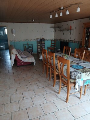 Un coin repas avec une longue table, des chaises et une nappe à motifs. On y trouve une bibliothèque et un fauteuil recouvert d’un tissu à fleurs.
