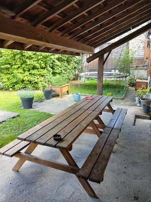 Una mesa de picnic de madera con bancos bajo una pérgola de madera, rodeada de plantas en macetas y un jardín.