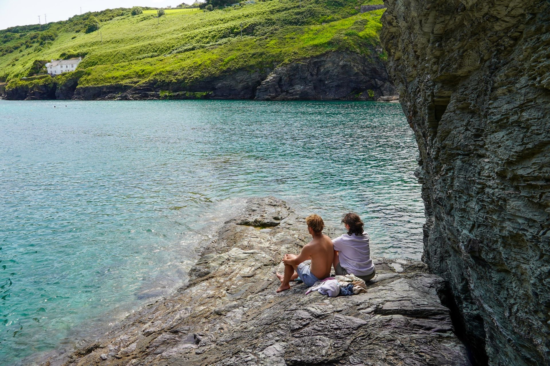 Two people sitting on a rocky cliff overlooking a body of water with a house in the background.