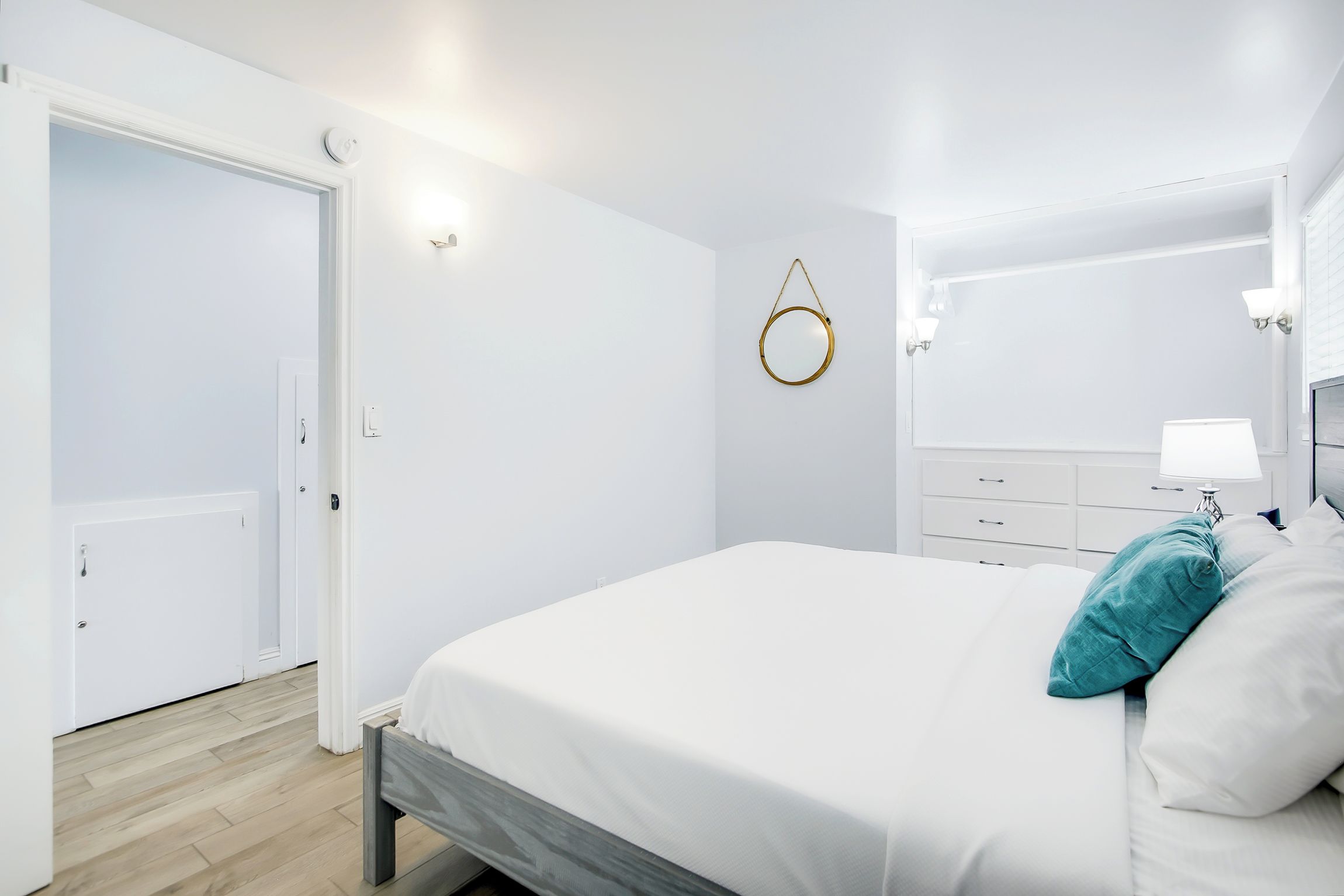 Une chambre à coucher avec un lit, une table de chevet avec une lampe, un miroir rond fixé au mur et une porte donnant sur une autre pièce.