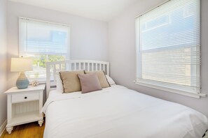 A bedroom with a white bed, a nightstand with a lamp, and two windows with blinds. - Perfect For Families, Walk To Beach & Shops! (Newport Beach)