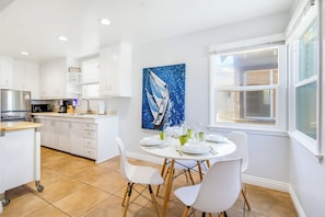 A modern kitchen with white cabinets, a stainless steel refrigerator, and a dining area with a round table and white chairs. - Perfect For Families, Walk To Beach & Shops! (Newport Beach)