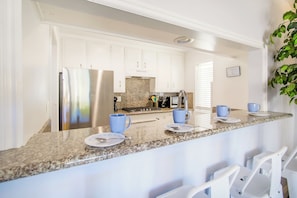 A kitchen with white cabinets, a stainless steel refrigerator, a granite countertop, and a bar area with blue cups and plates. - Short Walk To Sand Prefect For Families, Lrg Patio (Newport Beach)