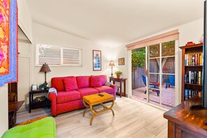 Ein Wohnzimmer mit einem roten Sofa, einem Holztisch, einem Bücherregal und einer Glastür, die zu einem Außenbereich führt.