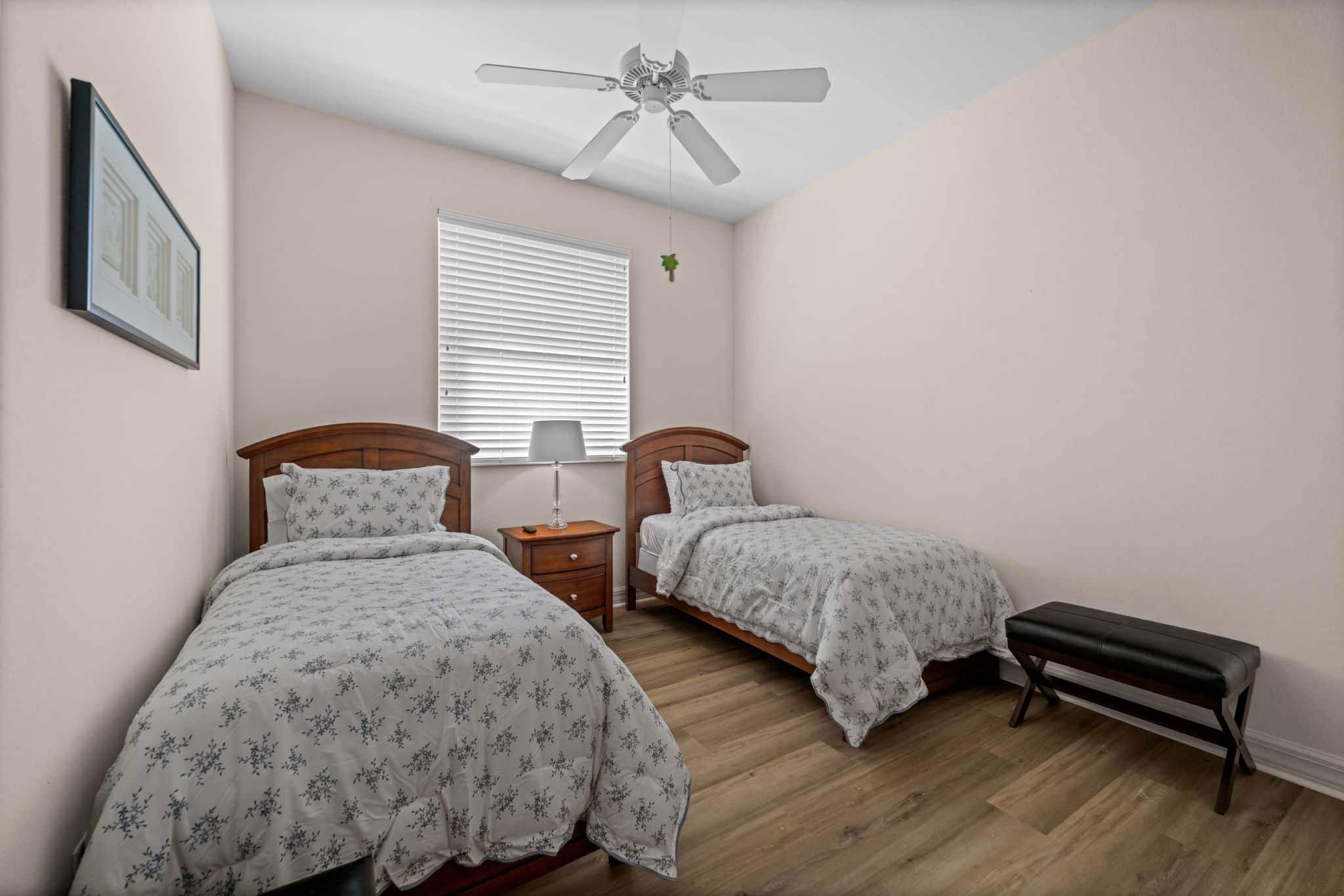 A bedroom with two beds, a bench, a ceiling fan, a window with blinds, and a framed picture on the wall.