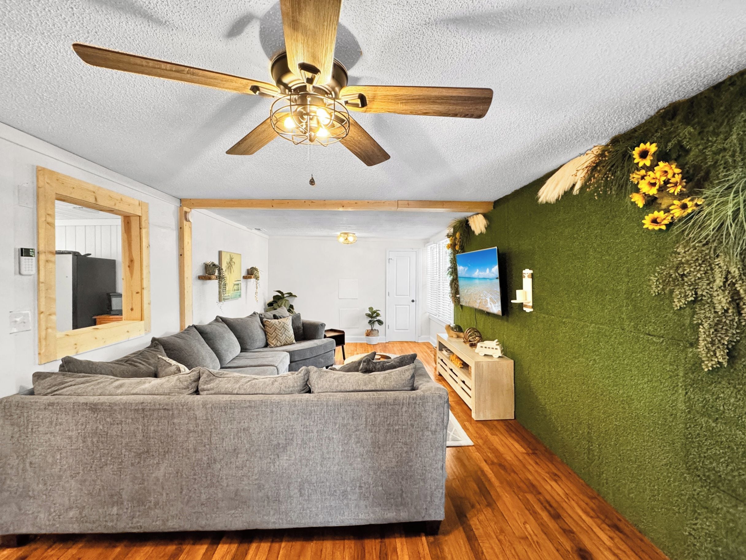 Una sala moderna con un sofá seccional gris, ventilador de techo de madera y una pared verde con un televisor montado.