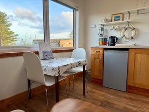A kitchen with a dining table, chairs, a refrigerator, and a window with a view of buildings and trees. - Seaview One Bed Annexe - Pets Welcome - Parking (Isle of Lewis)