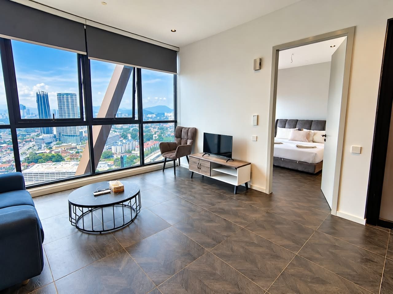 Una sala de estar moderna con un ventanal grande, un sofá, una mesa de centro, un televisor, y un dormitorio con una cama y una mesita de noche.