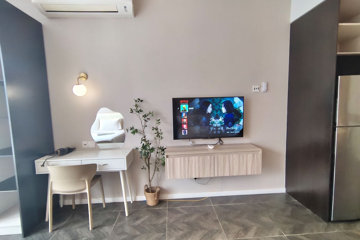A modern living room with a flat-screen TV mounted on the wall, a white desk with a chair, a potted plant, and a built-in wooden cabinet.