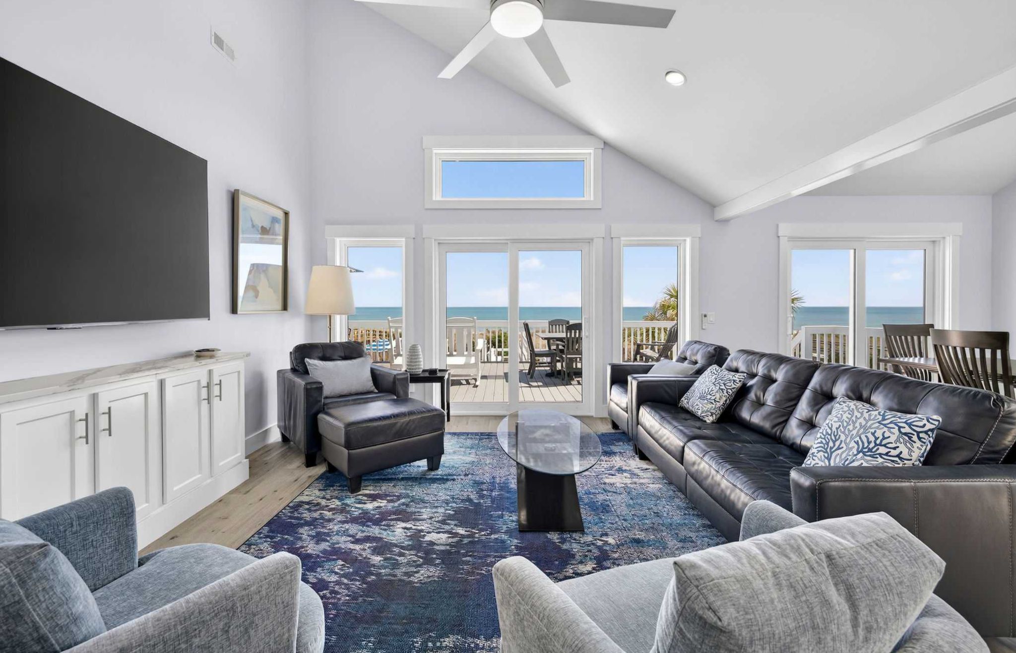 A spacious living room with a large flat-screen TV, comfortable seating, and a view of the ocean through large windows.