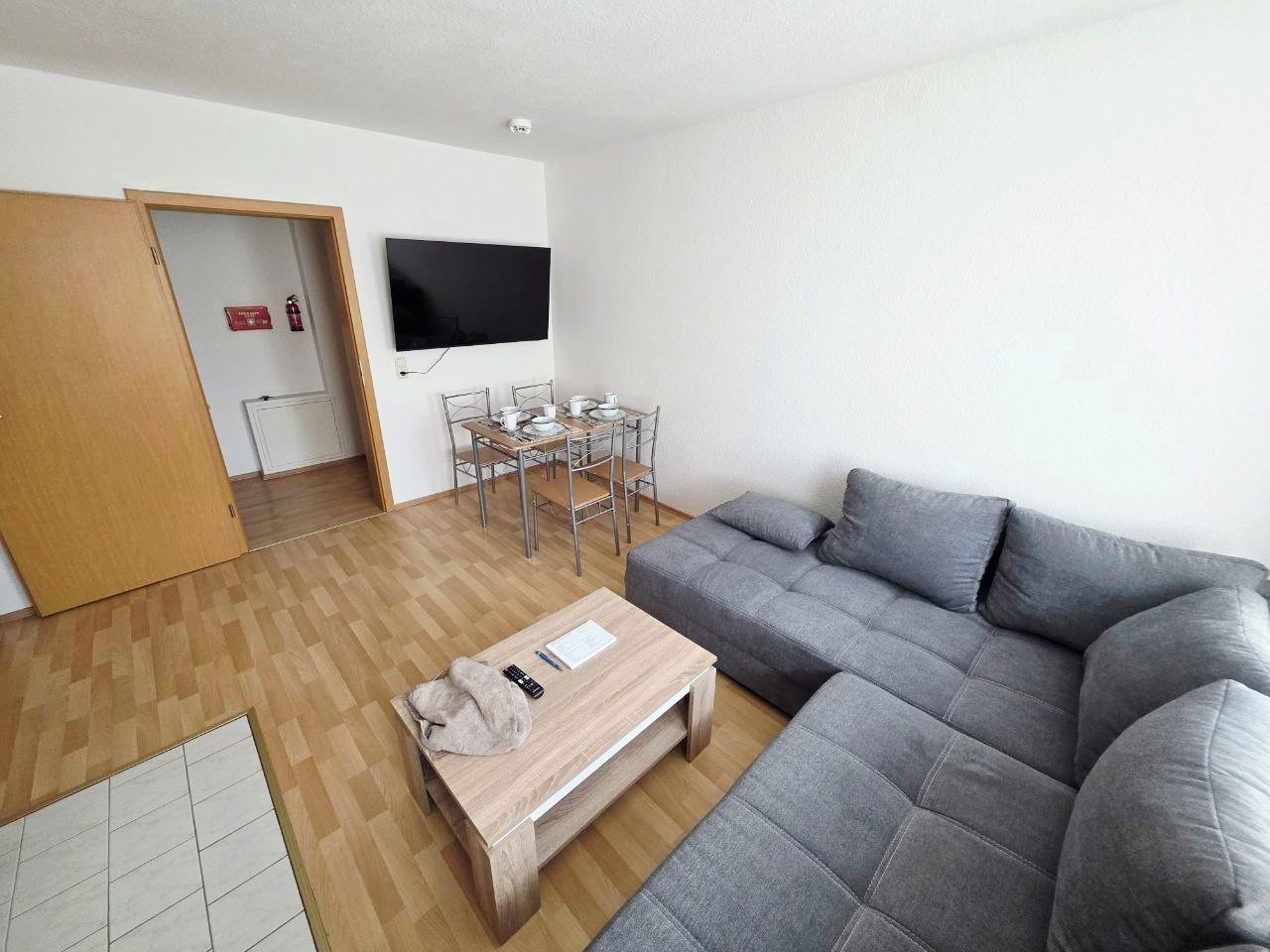 A modern living room with a grey sectional sofa, a wooden coffee table, a TV mounted on the wall, and a dining area with a table and chairs.