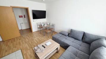A modern living room with a grey sectional sofa, a wooden coffee table, a TV mounted on the wall, and a dining area with a table and chairs.