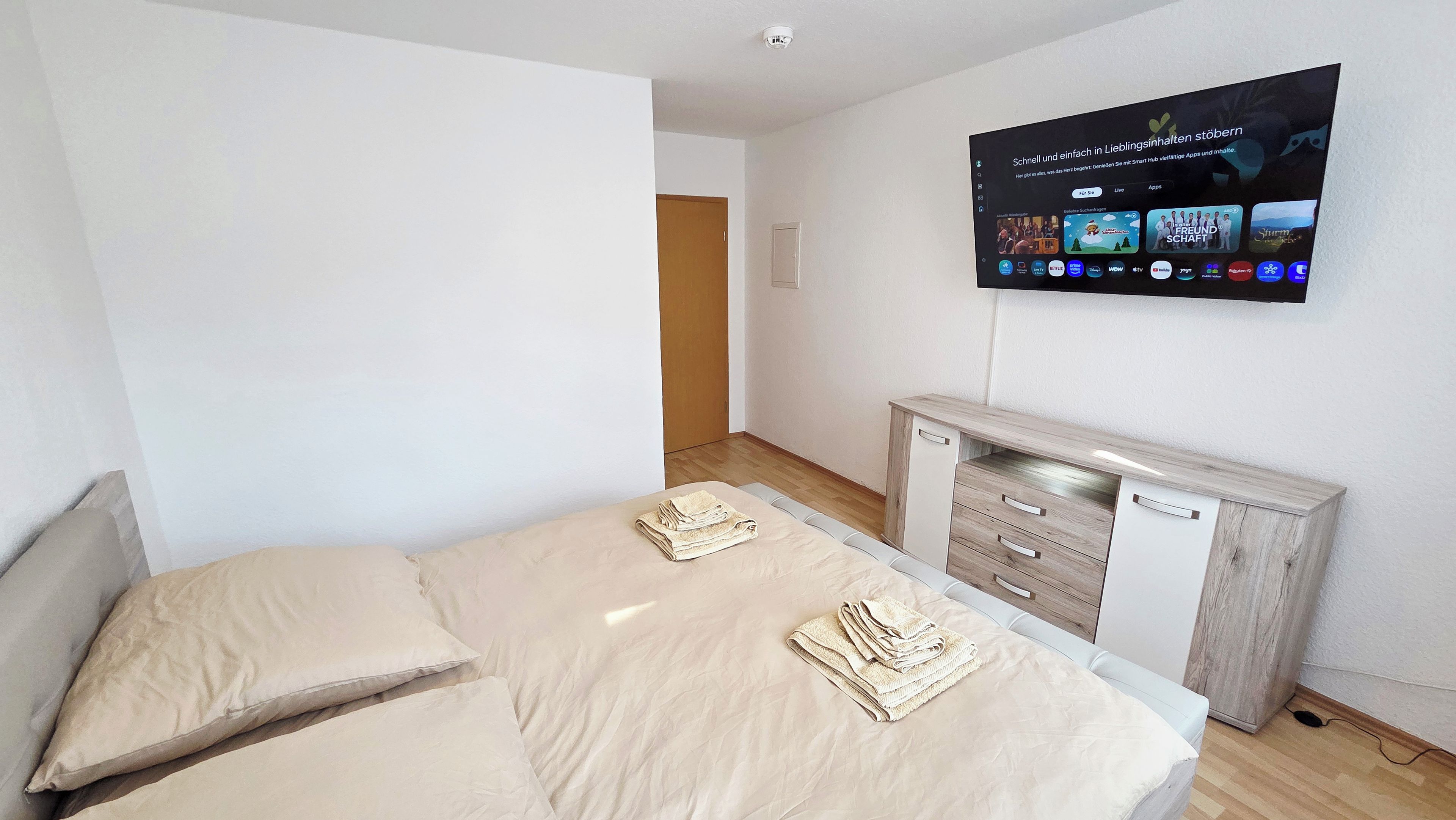 Une chambre à coucher comprenant un lit, une table de chevet, un téléviseur fixé au mur et une porte donnant sur une autre pièce.