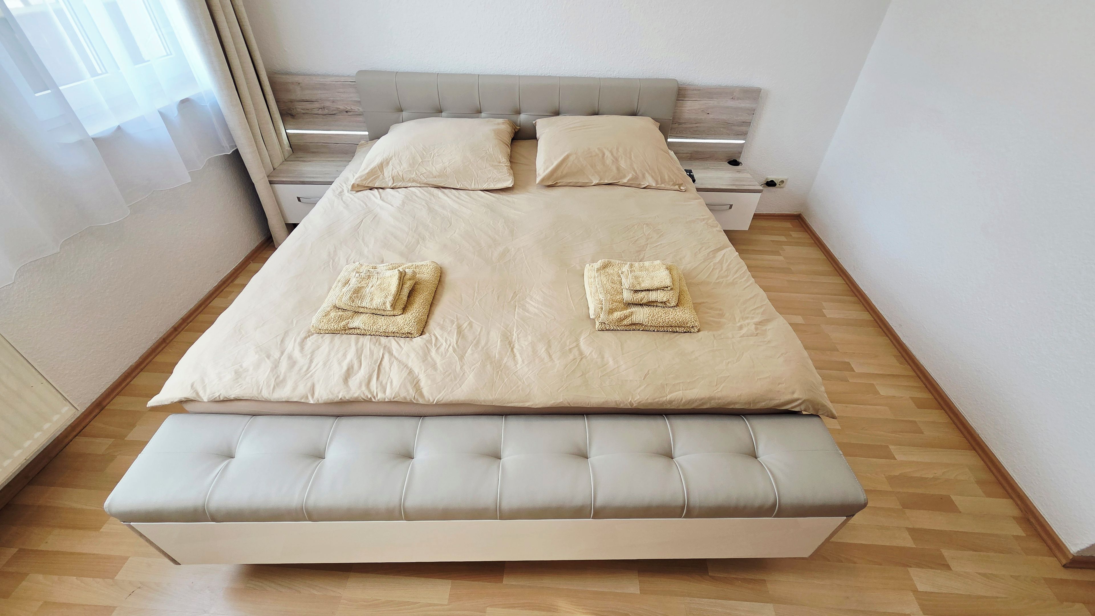Una cama bien hecha con cabecera, una mesita de noche y un banco al pie de la cama.