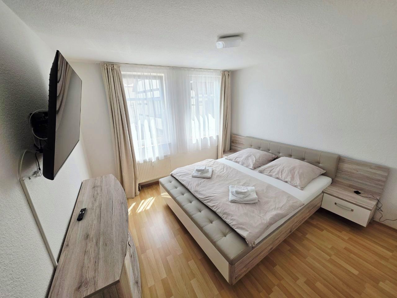 A bedroom with a bed, a bedside table, a TV mounted on the wall, and a window with curtains.