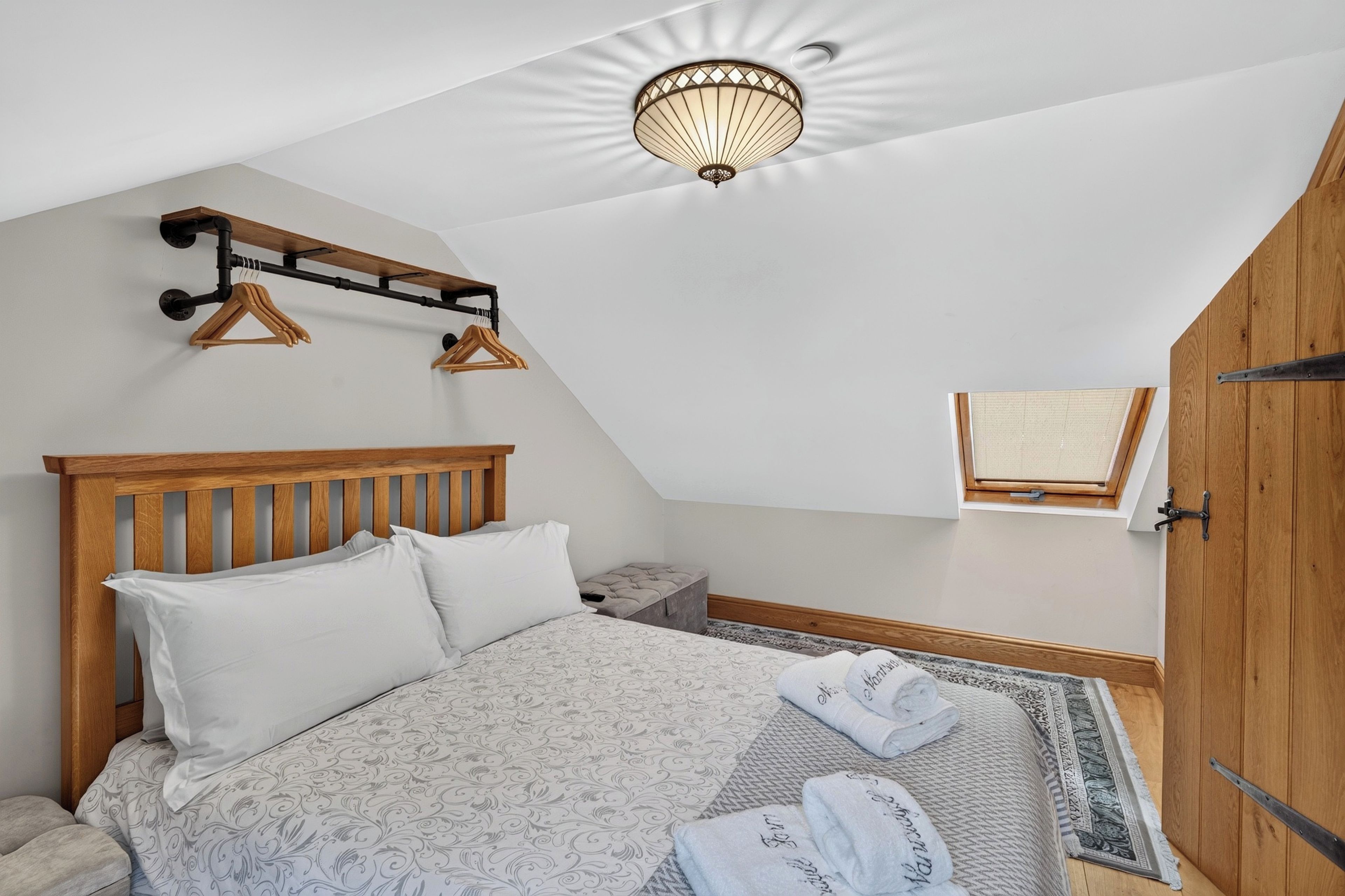 A bedroom with a sloped ceiling, a wooden bed frame, a bedside table, a hanging light fixture, and a small window.