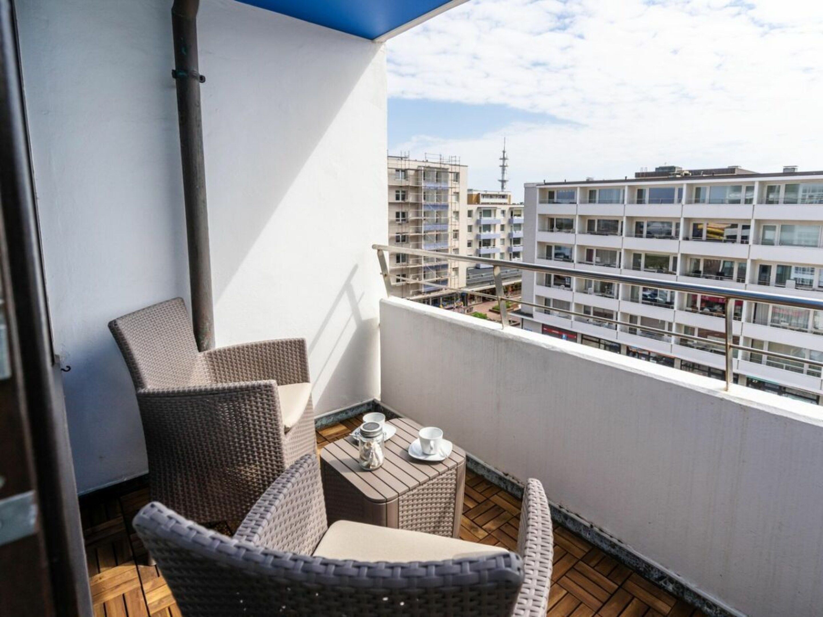 A balcony with wicker furniture, a small table with a cup and saucer, and a view of a cityscape with buildings.