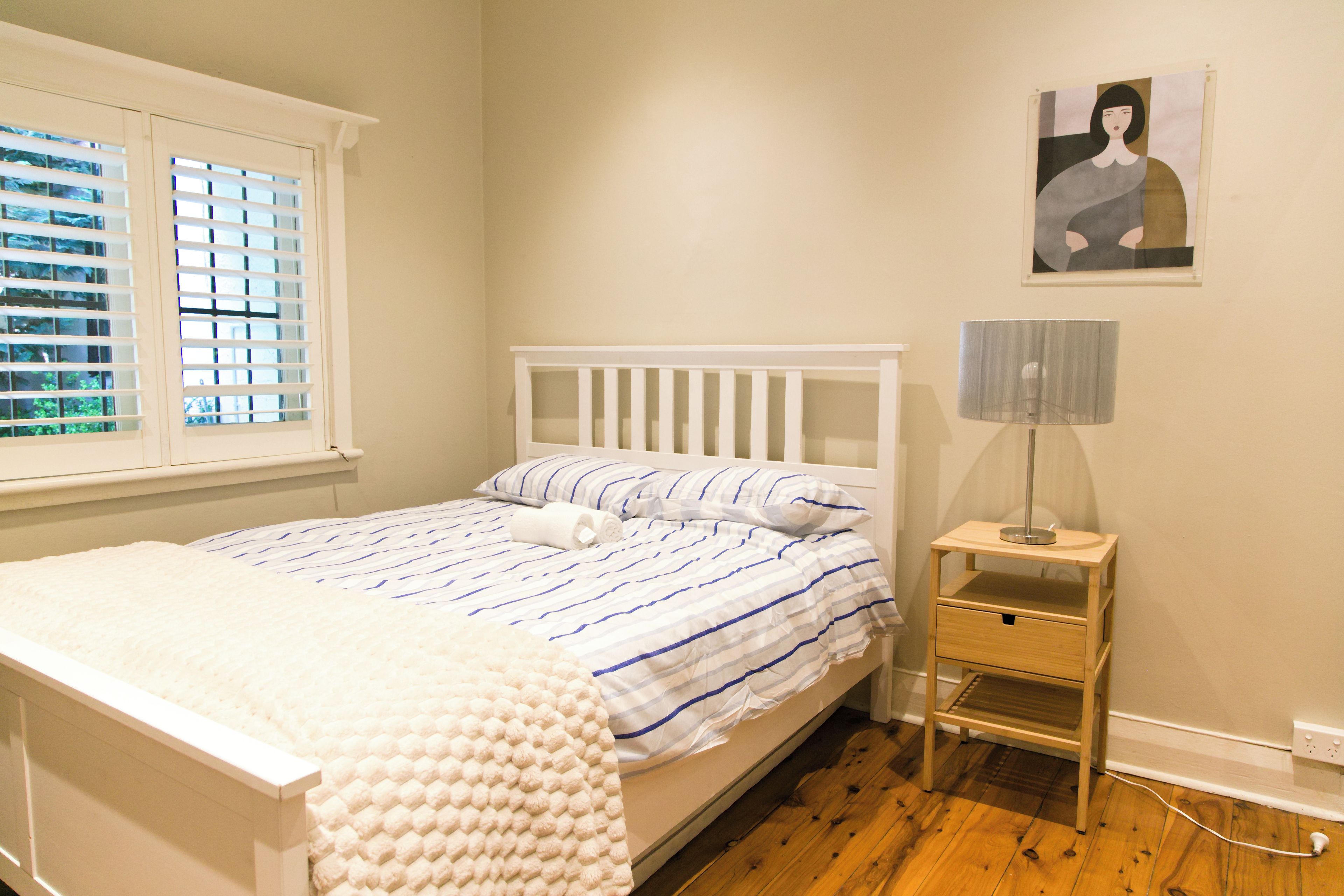 A bedroom with a bed, a bedside table with a lamp, a window with blinds, and a wall-mounted picture.