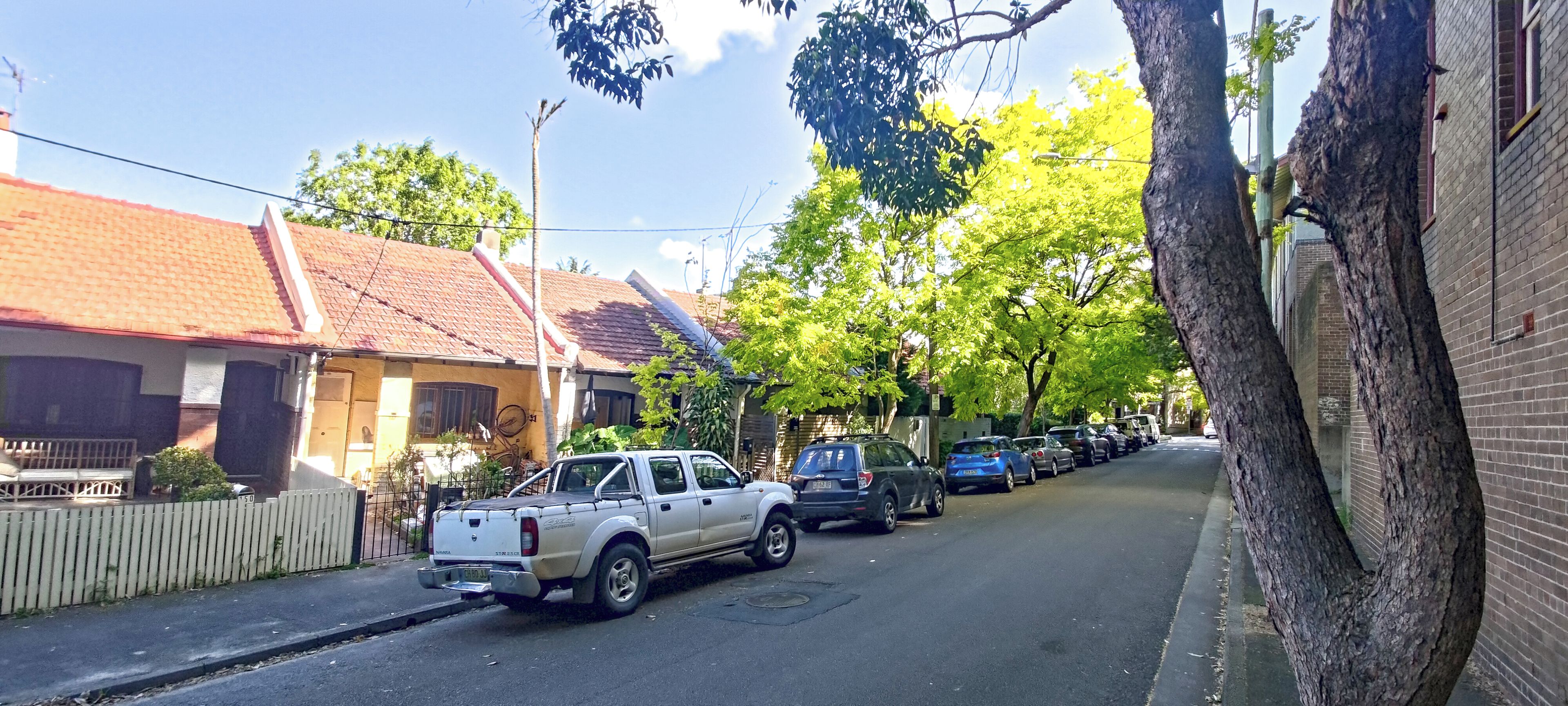 A residential street with parked cars, houses with red-tiled roofs, and trees.