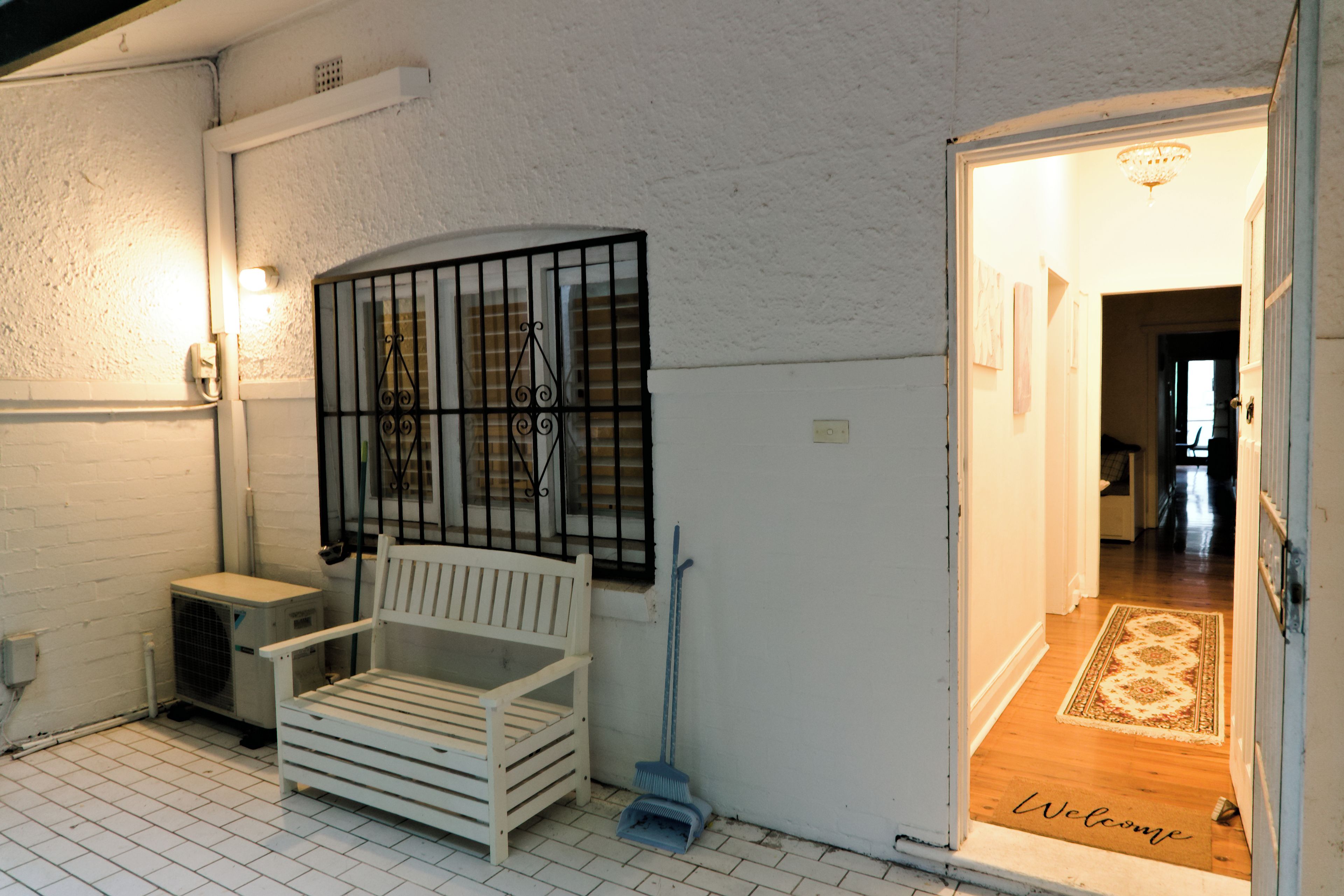Un banc blanc avec une rambarde en métal, un balai et un paillasson devant l’entrée d’un bâtiment aux murs blancs et au sol carrelé.