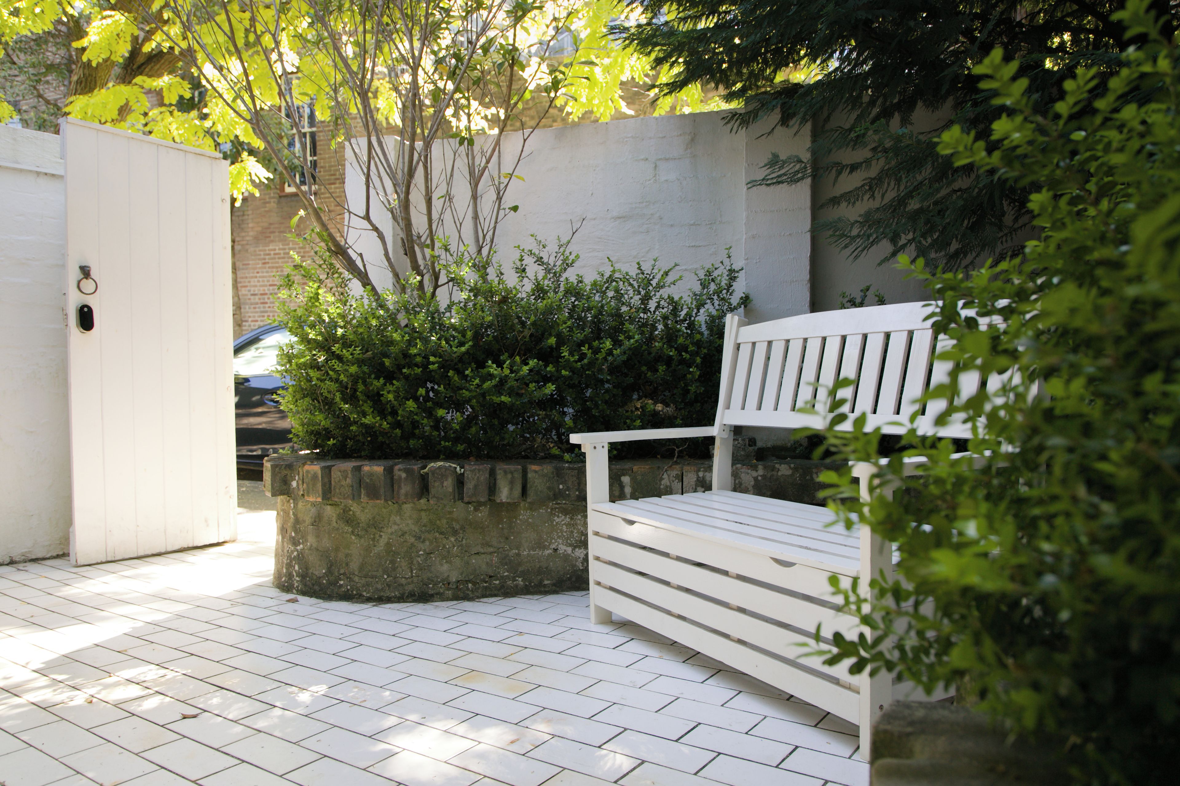 A white bench in a garden area with a paved walkway, surrounded by greenery and a white wall.